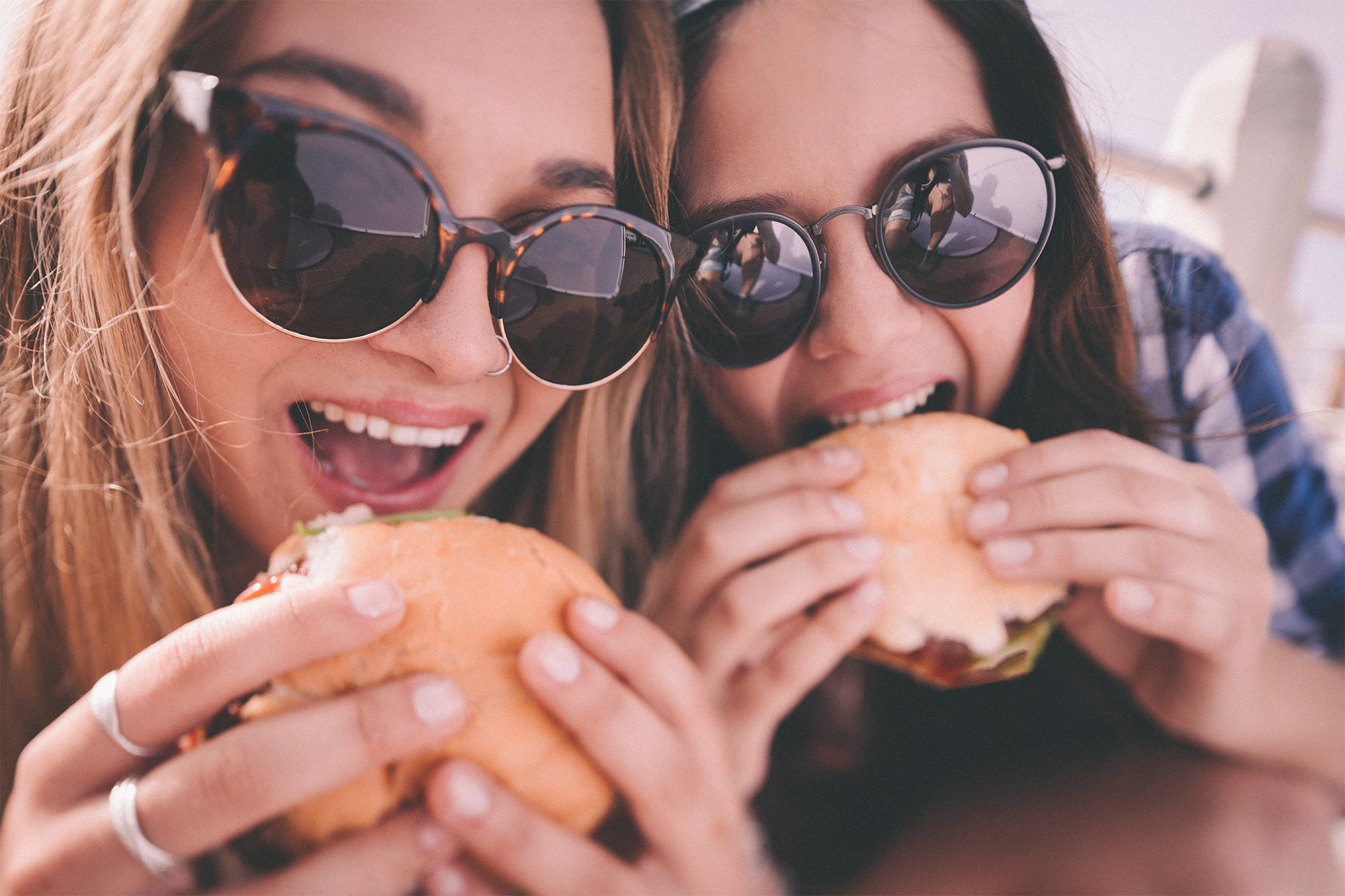 Closeup portrait of two teenage girls who are best friends, wearing hipster sunglasses about to take bites out of large burgers outdoors with a retro style develop
